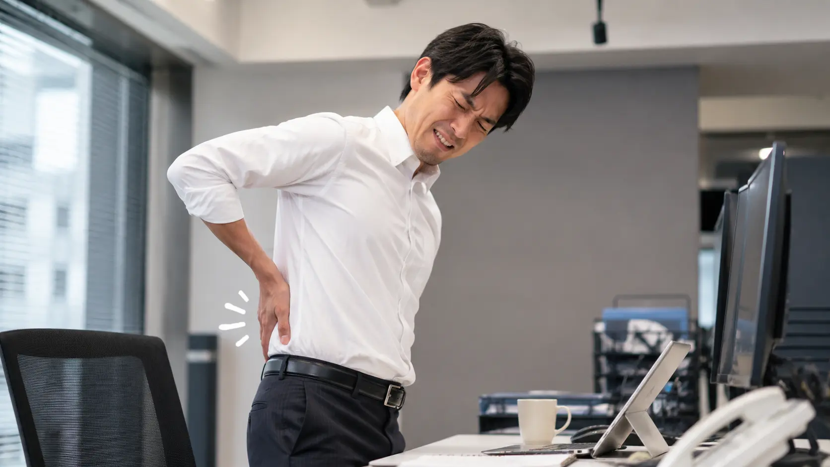 長時間のデスクワークや立ち仕事で、腰をトントンと叩きながら辛そうな表情をしているビジネスパーソン