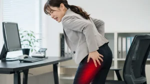 お尻から太ももの裏側にかけて、ピキッと走る痛みやしびれを辛そうに手で押さえているデスクワーカー